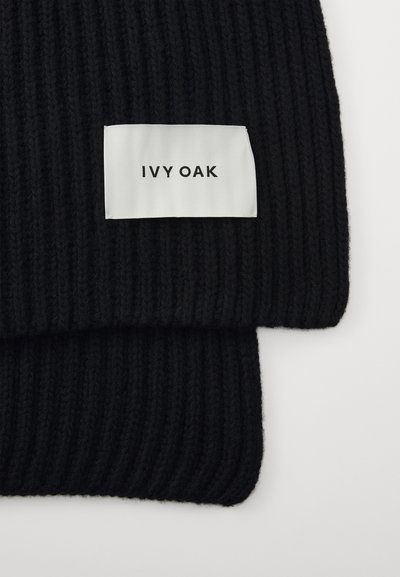 Melna ribota džinsa šalle ar baltu auduma uzlīmi, kurā melnā krāsā ir drukāts "IVY OAK". Tekstūra ir maiga un masīva.