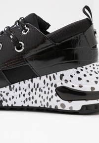 Primo piano di una sneaker nera con pelle testurizzata, suola spessa a macchie bianche e nere e lacci neri con accenti bianchi.