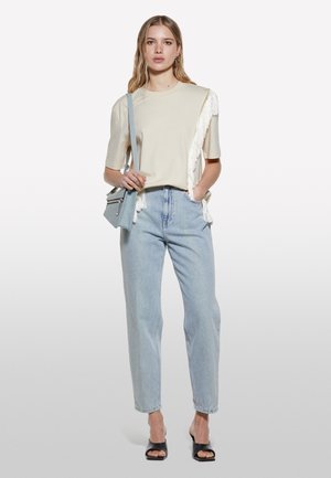 Beige T-Shirt mit Fransen-detail auf einer Seite, kombiniert mit hellblauen hochtaillierten Jeans. Schwarze Sandalen mit offenen Zehen und eine hellblaue Tasche.