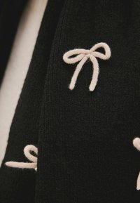 Sort strikvarer med små krèmefarvede broderede sløjfer, der tilføjer struktur og detaljer til stoffet.