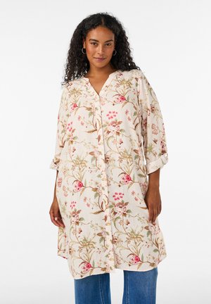 Vrouw met krullend haar die creolen draagt en een lange crèmekleurige bloemenjurk met knopen over een blauwe spijkerbroek, staande tegen een witte achtergrond.