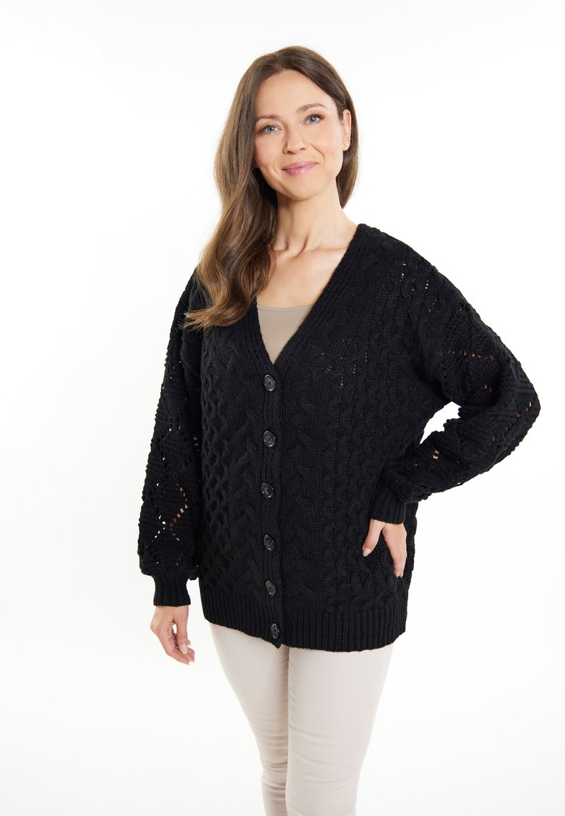 Cardigan nero in maglia con scollatura a V e chiusura a bottoni, caratterizzato da motivi testurizzati e polsini a costine. Indossato sopra una maglietta beige.