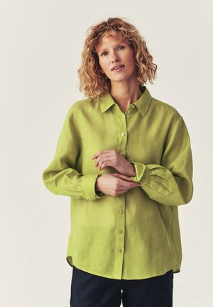 Frau mit lockigem Haar, die ein locker sitzendes hellgrünes Hemd mit Knöpfen trägt und ihren Ärmel justiert, vor einem einfachen hellen Hintergrund stehend.