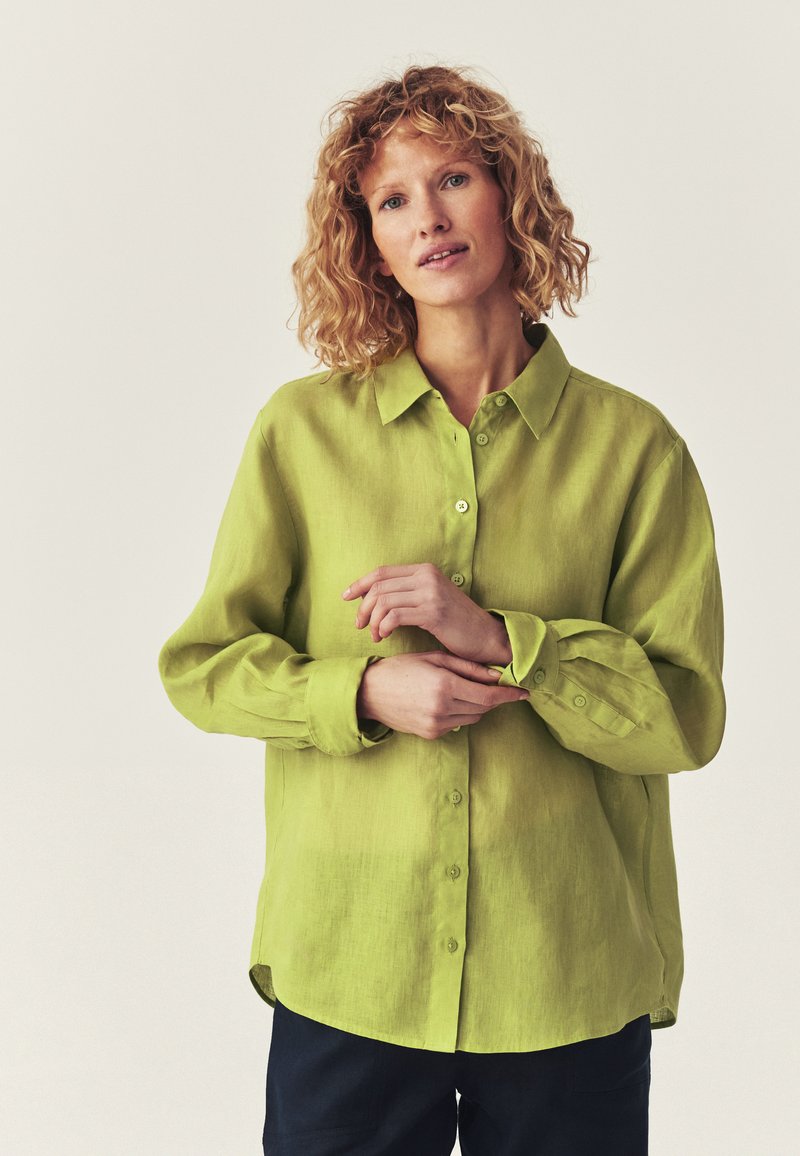 Femme aux cheveux bouclés portant une chemise ample boutonnée vert vif, ajustant sa manchette, debout devant un fond clair uni.