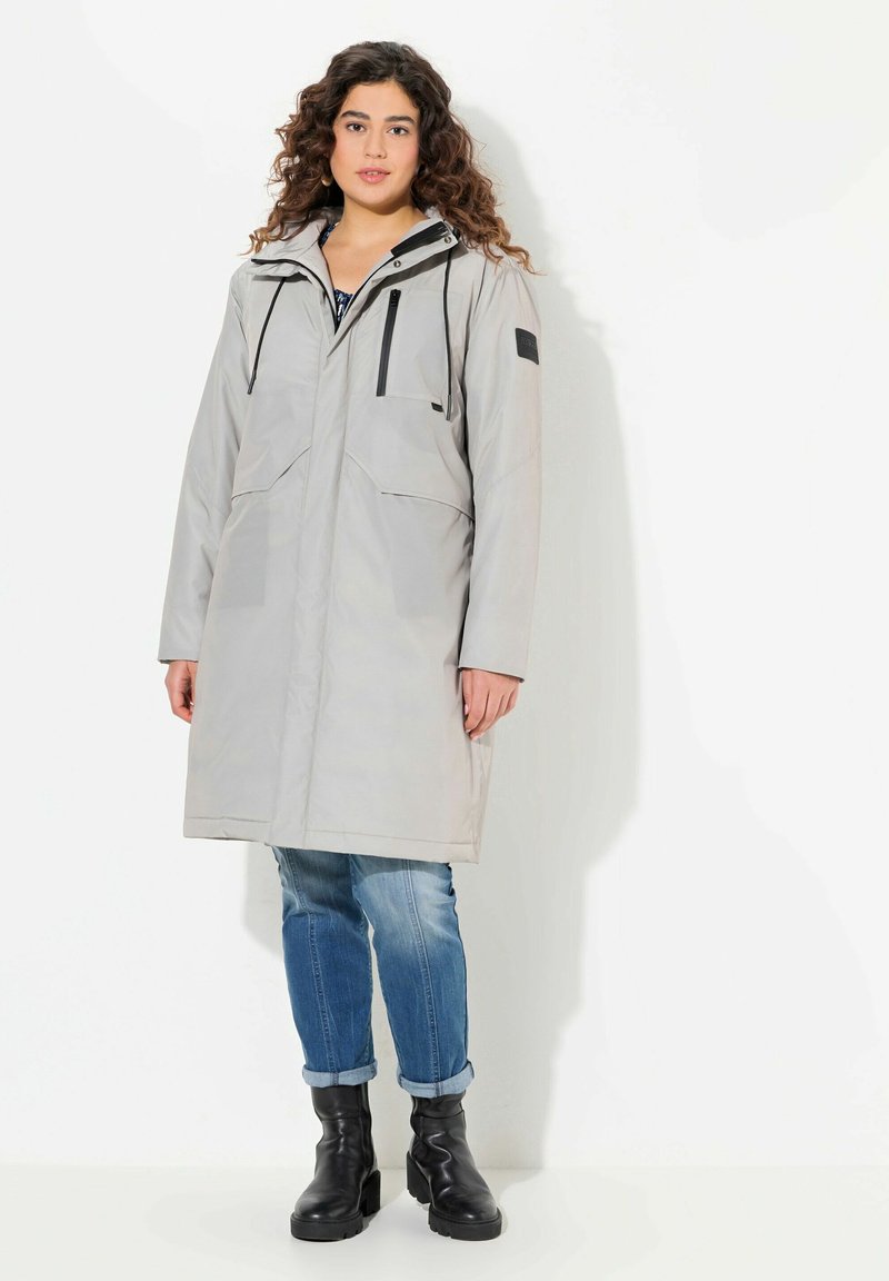 Cappotto grigio chiaro fino al ginocchio con cappuccio, due grandi tasche frontali, cerniere nere e un tessuto dalla texture interessante. Abbinato a jeans blu con risvolti e stivali neri.