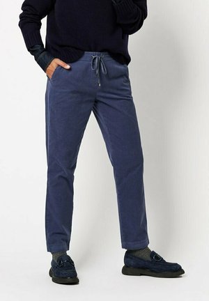 Person trägt marineblaue Kordelzughose, dunkle Slipper, graue Socken und einen dunklen Pullover, steht mit einer Hand in der Tasche.