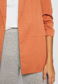 Lekka, rdzawo-pomarańczowa marynarka o luźnym kroju i rękawach do łokci, zestawiona z teksturowanymi, szarymi spodniami w wzory.