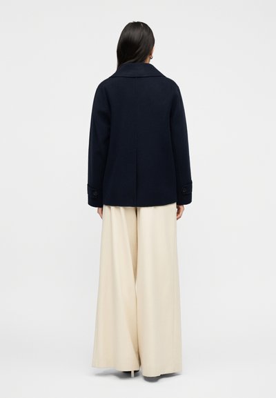 Γυναίκα με μακριά σκούρα μαλλιά, ντυμένη με σκούρο μπλε παλτό και εκρού παντελόνια φαρδιάς γραμμής, στέκεται γυρισμένη πλάτη σε ένα απλό φόντο.