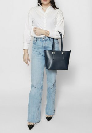 Donna con camicetta bianca e jeans azzurri, che tiene una borsa tote blu scuro, in piedi su sfondo bianco uniforme.