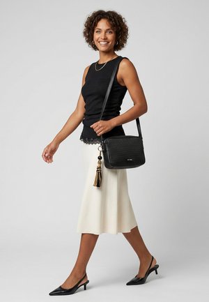 Femme marchant portant un haut noir sans manches, une jupe midi blanche, des talons slingback noirs, et portant un sac à épaule noir texturé avec un pompon.