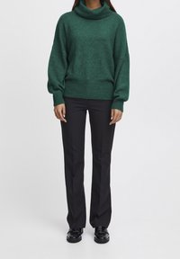 Maglione verde lavorato a maglia con colletto alto, vestibilità morbida e bordi a costine abbinato a pantaloni neri aderenti e scarpe lucide.