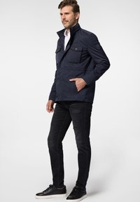 Granatowa kurtka z wysokim kołnierzem, dwoma przednimi kieszeniami i zapinanymi detalami, w połączeniu z czarnymi przetartymi dżinsami i ciemnymi sneakersami.