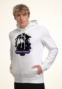 Uomo biondo che indossa un hoodie bianco con una grafica nera e viola raffigurante una donna, un corvo, dei fiori e il testo "Socialmente Distanziati", con le mani nelle tasche.