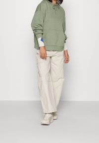 Sweat à capuche vert avec une poche frontale et des manches crème ornées de touches bleues, associé à un pantalon cargo beige et des baskets de couleur claire.