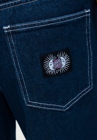 Πίσω τσέπη σκούρου μπλε τζιν με λευκή ραφή και μαύρο ορθογώνιο έμβλημα που απεικονίζει έναν ήλιο και φεγγάρι, με το κείμενο «Santa Cruz».