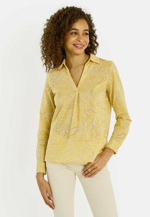 Femme aux cheveux bouclés portant un chemisier jaune à manches longues avec un motif œillet en forme de feuille et un pantalon beige clair, souriante et regardant sur le côté.