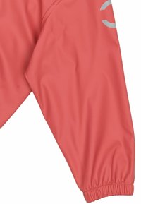 Manche d'une veste imperméable rouge avec poignet élastique et logo réfléchissant partiel sur le haut du bras, sur fond blanc.