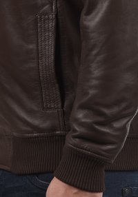 Donkerbruin leren jack met details van mouw en zijzak over blauwe denim jeans, met zichtbare geribbelde manchetten en textuurstiksels.