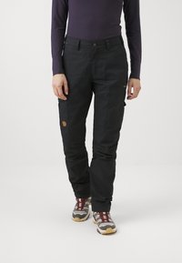 Melni kravas bikses ar vairākiem kabatām, pieguļošs dizains un smalka teksturēta apdare. Valkātas ar pelēkiem un sarkaniem pārgājienu apaviem.