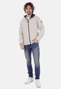 Chaqueta ligera de color gris claro con capucha y detalles en negro, con una textura suave. Combinada con jeans azules y zapatillas blancas.