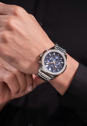 Polso maschile con orologio da uomo in metallo argento con quadrante cronografo blu e multiple lancette secondarie, mentre si regola l'orologio su sfondo di camicia nera a maniche lunghe.
