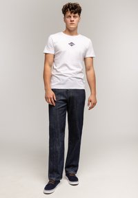 Młody mężczyzna stoi w białym t-shircie z grafiką, ciemnoniebieskich dżinsach i granatowych trampkach na tle jasnej, jednolitej ściany.