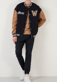 Marinblå och beige varsityjacka med "W"-logotyp och texten "Classy", randiga ribbade ärmslut och krage, bärs med beige skjorta och mörka byxor.