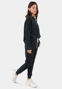 Donkergroene sweatshirt en bijpassende joggingbroek gemaakt van zacht materiaal, met een relaxte pasvorm, elastische boorden en een taille met trekkoord. Draagt met witte sneakers.