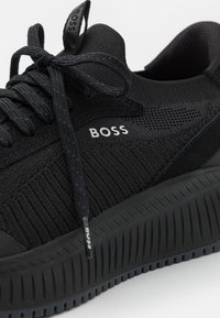 Svarta sneakers med en texturerad stickad ovandel, tonade snören, en märkesetikett och en tjock, räfflad gummisula för bättre grepp.