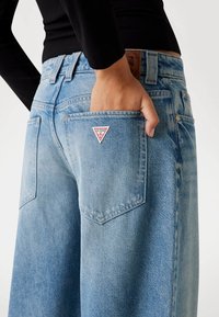 Jeans in denim azzurro chiaro con una vestibilità ampia, caratterizzati da un patch a forma di triangolo, tasche posteriori e una finitura texture. Bottoni e vita visibili.