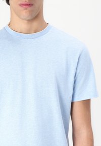 Lys blå bomull T-skjorte med korte ermer, rund hals og en liten logo på brystet. Stoffet ser mykt og glatt ut.
