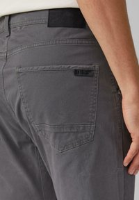 Pantalons en coton gris avec une poche arrière, présentant des coutures discrètes et une étiquette en tissu. La texture semble lisse et la coupe est ajustée.