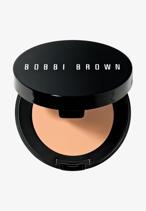 Kompaktowy puder w czarnym okrągłym opakowaniu z białym napisem „BOBBI BROWN”. W środku znajduje się gładka, jasnobeżowa powierzchnia produktu.