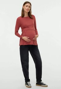 Terhes nő áll, a hasát fogja, hosszú ujjú rozsdabarna felsőt, laza fekete nadrágot és fehér fűzős fekete tornacipőt visel.