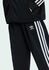 Černé teplákové soupravy se třemi bílými pruhy po stranách. Materiál vypadá hladce a má detail se zipovou kapsou. Viditelné logo Adidas.