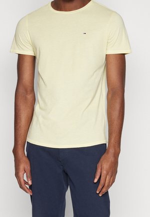 Mężczyzna ubrany w prosty jasnofioletowy T-shirt z krótkim rękawem z małym logo na piersi i ciemnoniebieskie spodnie, stojący na neutralnym tle.