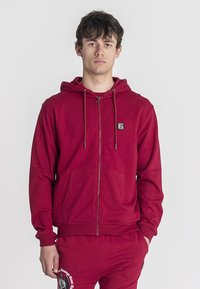 Červená mikina na zips s kapucňou na stiahnutie, prednými vreckami a nášivkou loga na hrudi. Jej tkanina vyzerá mäkko s jemnou textúrou.