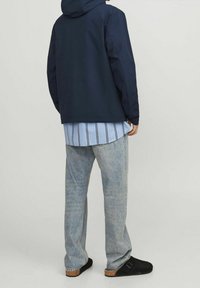Persona che indossa una giacca blu navy, una camicia a righe azzurro chiaro, jeans sbiaditi e zoccoli neri senza lacci, in piedi davanti a uno sfondo semplice.