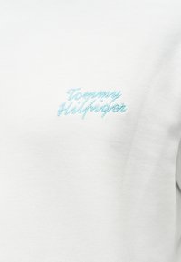Λευκό ύφασμα με ελαφρώς γαλάζιο κείμενο «Tommy Hilfiger» κεντημένο με καλλιγραφική γραφή στο ύψος του στήθους.