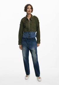 Olijfgroene korte jas met rits en schouderepauletten, gecombineerd met blauwe denim broeken met diverse zakken en een losse pasvorm.