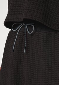 Tissu noir texturé avec un motif nid d'abeille, comportant une taille à cordon de serrage avec des accents bleus contrastants et des embouts noirs.