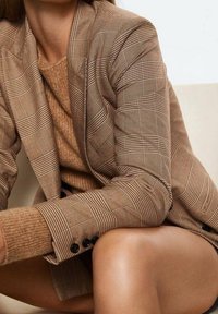 Brązowa kraciasta marynarka z wyciętym klapą, pod spodem teksturowany brązowy sweter oraz ciemne guziki na mankietach; widoczna gładka skóra.