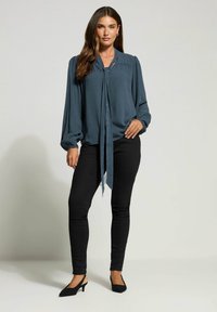 Mørk teal bluse med bindebånd i halsen, lange pufærmer og indsamlet detaljering. Sammen med sorte skinny jeans og sorte slingback sko.