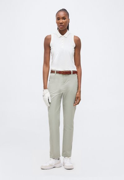 Femme portant un polo blanc sans manches, pantalon gris clair avec une ceinture marron, gant de golf blanc à la main gauche et baskets blanches, debout devant un fond blanc.