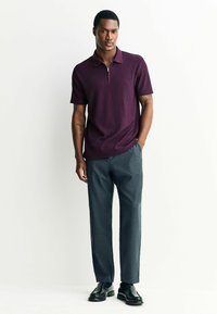 Polo en maille de couleur vin avec un col zippé, associé à un pantalon gris foncé ajusté et des chaussures noires, se tenant devant un fond neutre.
