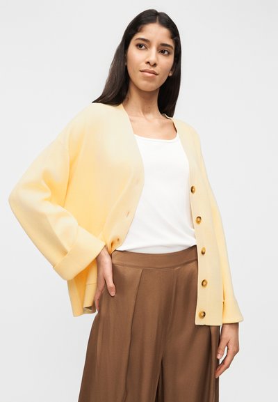 Γυναίκα που φοράει ανοιχτό κίτρινο ζακέτα με κουμπιά πάνω από λευκή φανέλα και φαρδιά καφέ παντελόνια, όρθια μπροστά σε απλό ανοιχτό φόντο.