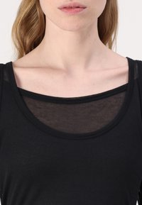 Černý bezrukávový top s kulatým výstřihem a průsvitným překrytím. Hladká textura a lehký materiál s přiléhavým střihem.