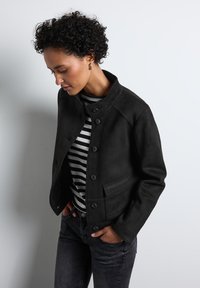 Svart mockajacka med hög krage, knappstängning och sidofickor, som bärs med en svart och vit randig skjorta samt mörka jeans.