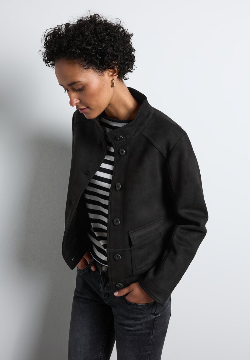 Svart mockajacka med hög krage, knappstängning och sidofickor, som bärs med en svart och vit randig skjorta samt mörka jeans.