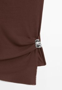 Brown fabric garment detail with a gathered side held by a small, irregularly shaped shiny silver metal clasp.